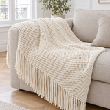 Plaid de canapé en maille ajourée crème à longues franges posé sur un canapé en lin beige
